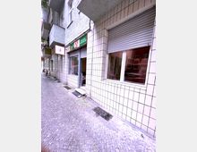 Eine schmale, gepflasterte Straße mit einem Wohn- und Geschäftshaus, dessen Fassade mit weißen Fliesen verkleidet ist. Im Erdgeschoss befinden sich Ladenräume mit Rolläden sowie ein geöffnetes Geschäft mit einer grünen Werbetafel.