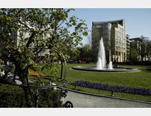 Ein Stadtpark mit einem zentralen Springbrunnen, umgeben von einer Grünfläche, Blumenbeeten und Bäumen. Im Hintergrund sind moderne mehrstöckige Gebäude sichtbar, während im Vordergrund ein Baum und ein Kinderwagen zu sehen sind.