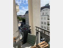 Ein kleiner Balkon in einem Wohngebäude mit einer gepolsterten Sitzfläche und einem Holztisch im Vordergrund. Der Blick zeigt ein städtisches Wohnviertel mit weißen, mehrstöckigen Gebäuden, teilweise mit schrägen Dächern, und einer Straße mit parkenden Autos sowie Bäumen.