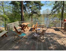 Eine Holzterrasse im Freien, umgeben von Bäumen und mit Blick auf einen See. Auf der Terrasse befinden sich Holztische, -bänke und Liegestühle, mit einem Geländer aus Netzmaterial als Absicherung.