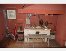 Historisches Kücheninterieur mit gemauertem Ofen, Holzscheiten und diversen Küchenutensilien, darunter ein Samowar und Keramikgeschirr. Im Hintergrund hängen Werkzeuge und Schautafeln an einer rot gestrichenen Wand, rechts steht ein alter Webstuhl, links ein Feuerlöscher.