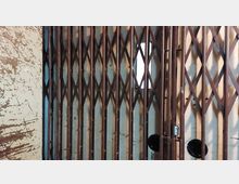 Metallisches Scherengitter einer alten Aufzugstür, mit rostbrauner Farbe und diagonalen Streben, vor einer beigen, abgenutzten Wand. Ein kleines Fenster im Hintergrund lässt Licht durch.