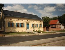 Ein einstöckiges gelbes Gebäude mit blauen Fensterläden und einer blauen Eingangstür steht an einer gepflasterten Straße. Im Hintergrund befindet sich ein traditionelles Gebäude mit rotem Ziegeldach und Fachwerkkonstruktion, umgeben von Bäumen.