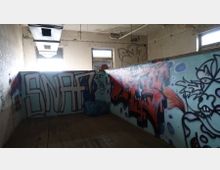 Ein verlassenes, heruntergekommenes Gebäude mit Wänden, die mit Graffiti bemalt sind, darunter Schriftzüge und abstrakte Zeichnungen. Der Raum hat hohe Fenster mit beschädigtem Glas und eine abblätternde Decke mit sichtbaren Rohren und Luftkanälen.