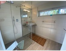 Modernes Badezimmer mit ebenerdiger Glasdusche, weißen Wandfliesen und grauem Bodenbelag. Rechts befindet sich ein Waschbecken mit Spiegel und ein schmales Fenster mit Blick ins Grüne, darunter ein Handtuchhalter.