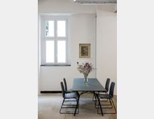 Ein minimalistischer Raum mit weißen Wänden, einem rechteckigen Tisch aus grauem Material und vier schwarzen Stühlen. Auf dem Tisch steht eine Vase mit trockenen Blumenzweigen, im Hintergrund ein großes Fenster und ein kleines Bild an der Wand.