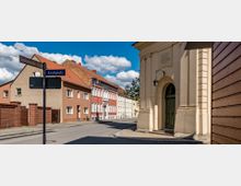 Straßenszene in einer Kleinstadt mit Häusern in verschiedenen Farben und Ziegeldächern; rechts im Vordergrund eine helle Kirchenfassade mit grünem Holztor und Lampe, links Straßenschilder mit den Aufschriften „Luckenwalder Straße“ und „Kirchplatz“.
