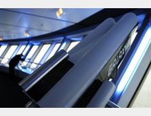 Ein modern gestalteter Innenraum mit großen, schrägen Fenstern, durch die helles Licht einfällt. Im Vordergrund ist ein futuristisches Modell mit der Aufschrift „GALAXY“ zu sehen, während im Hintergrund eine Person durch eines der Fenster schaut.