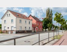 Zwei zweistöckige Wohngebäude, eines cremefarben und eines rot, mit Satteldächern und weißen Fenstern, stehen an einer Straße mit Gehweg und Bäumen. Am vorderen Gebäude ist ein Schild mit der Aufschrift „Boulevard Café“ angebracht, und Straßenlaternen mit Blumen hängen entlang der Straße.